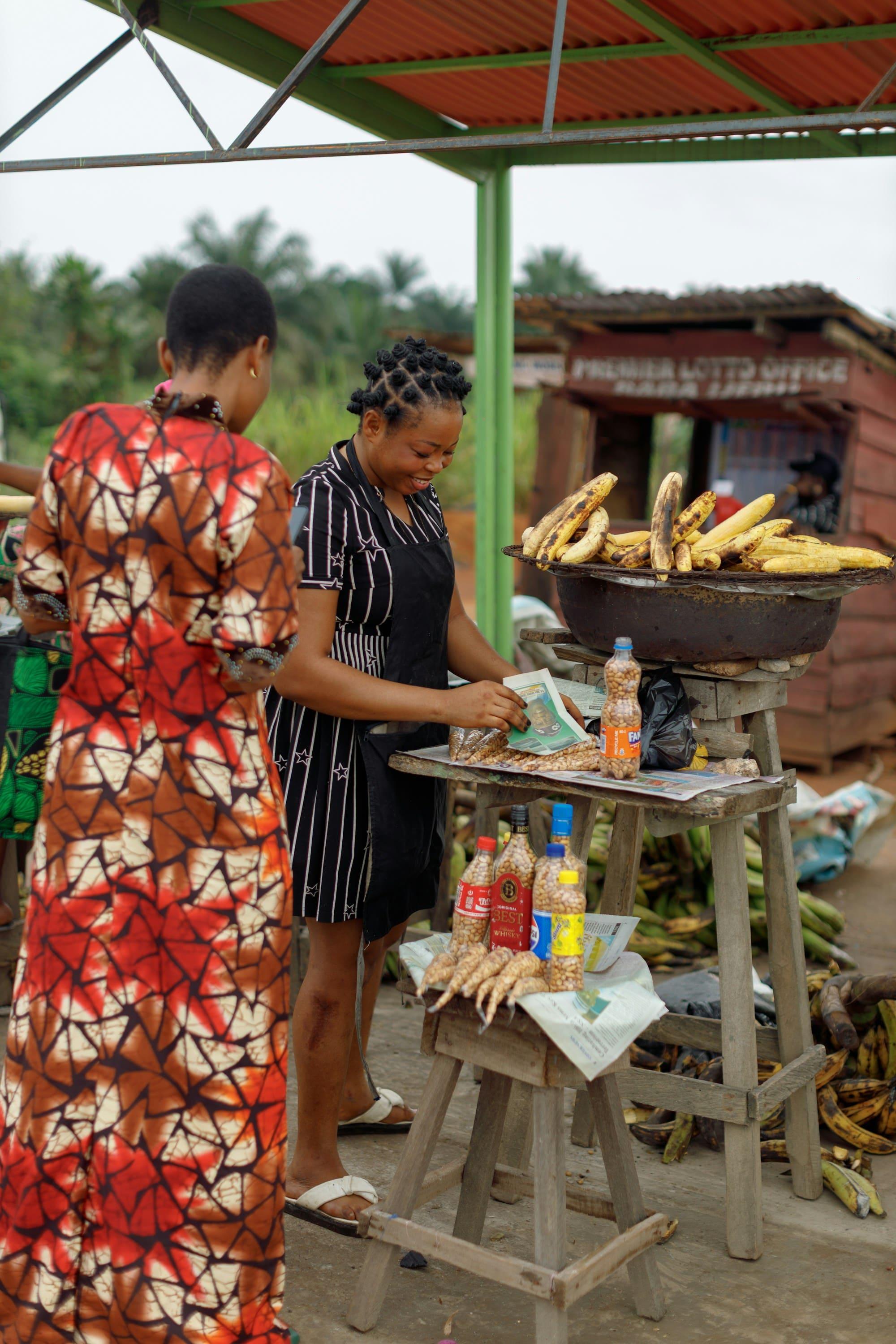 Two women talk at a roadside stall selling roasted plantain