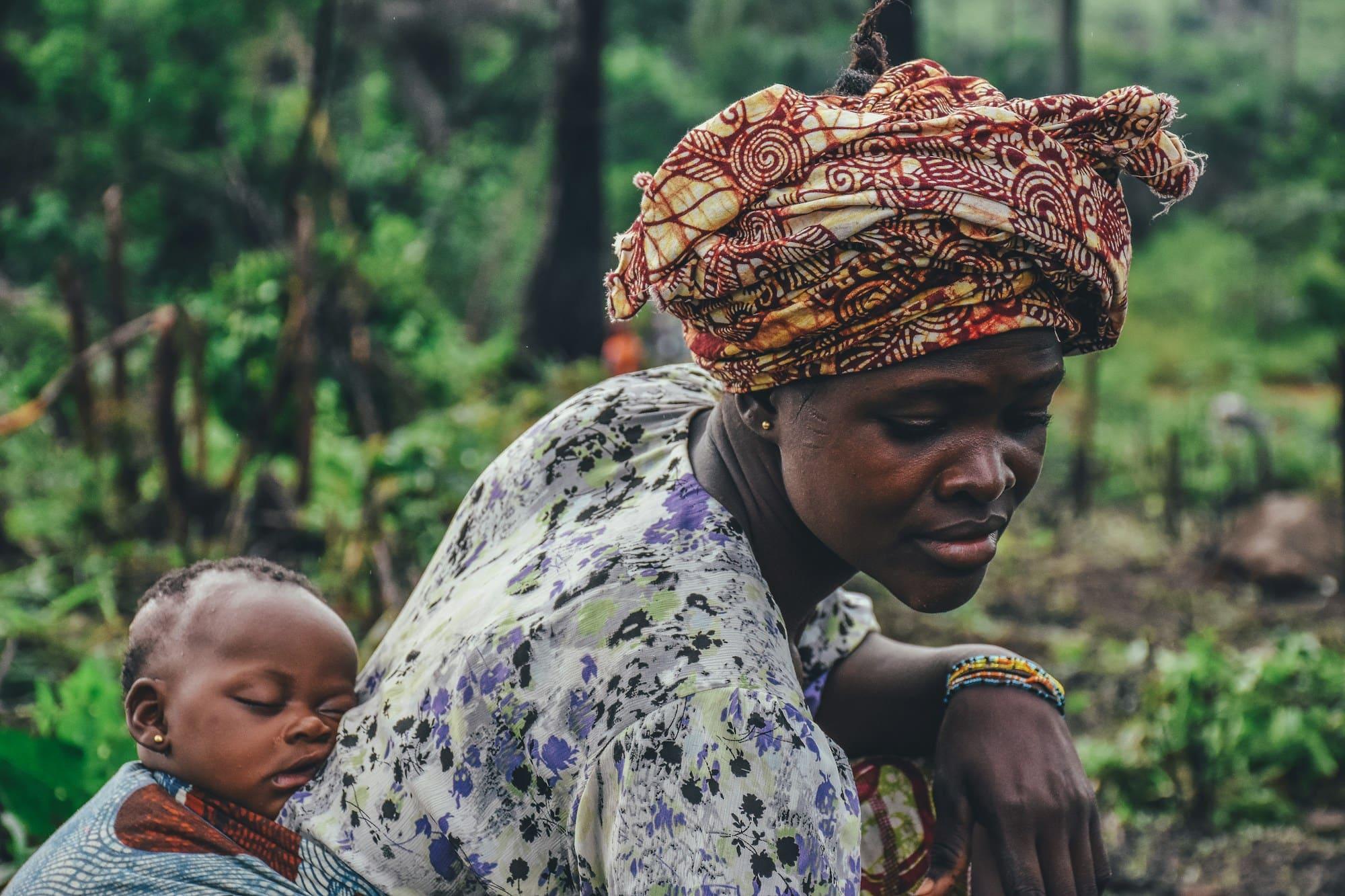 A woman in a patterned headwrap carries her sleeping baby on her back