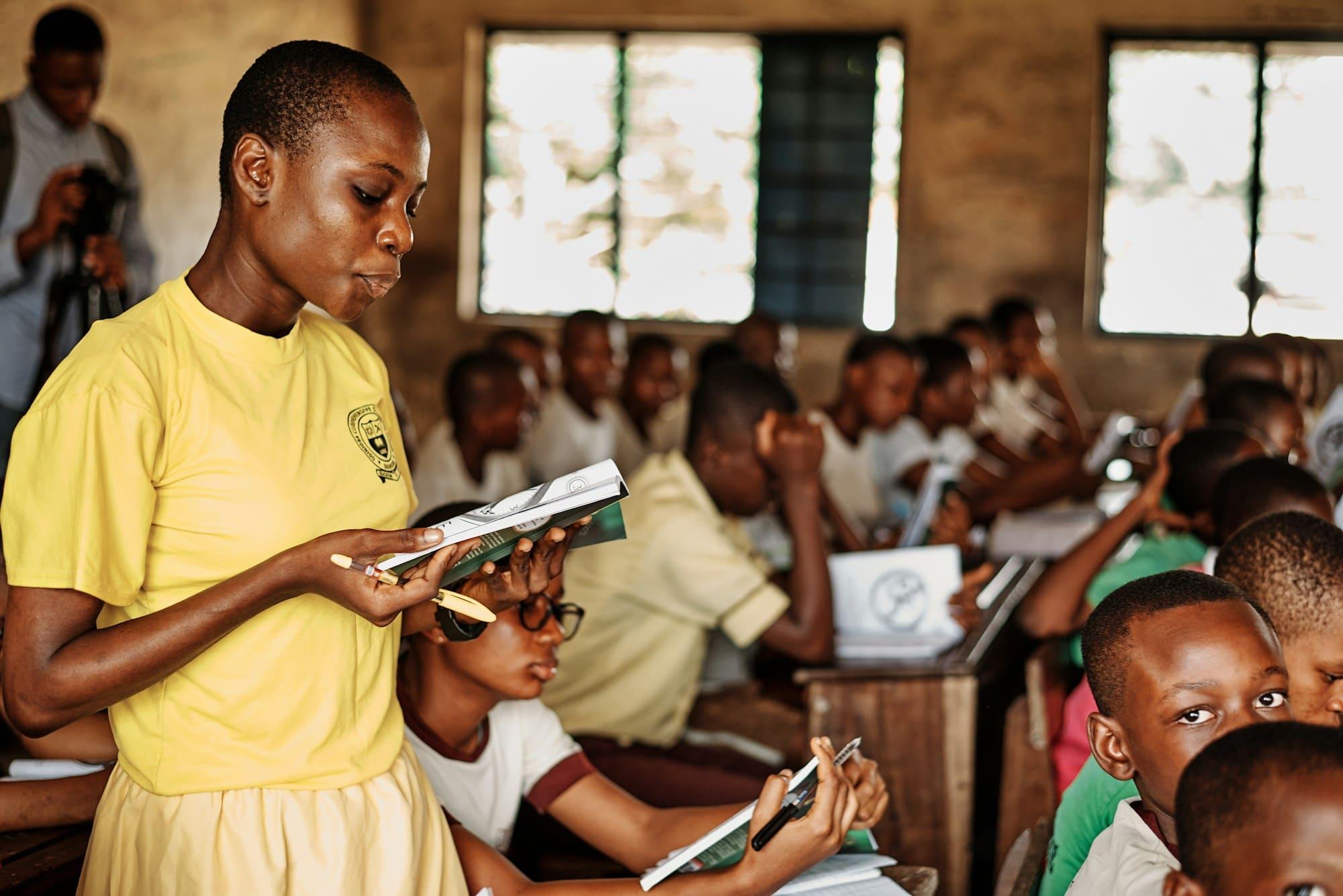 A young woman reads aloud from a book as pupils follow along at their desks