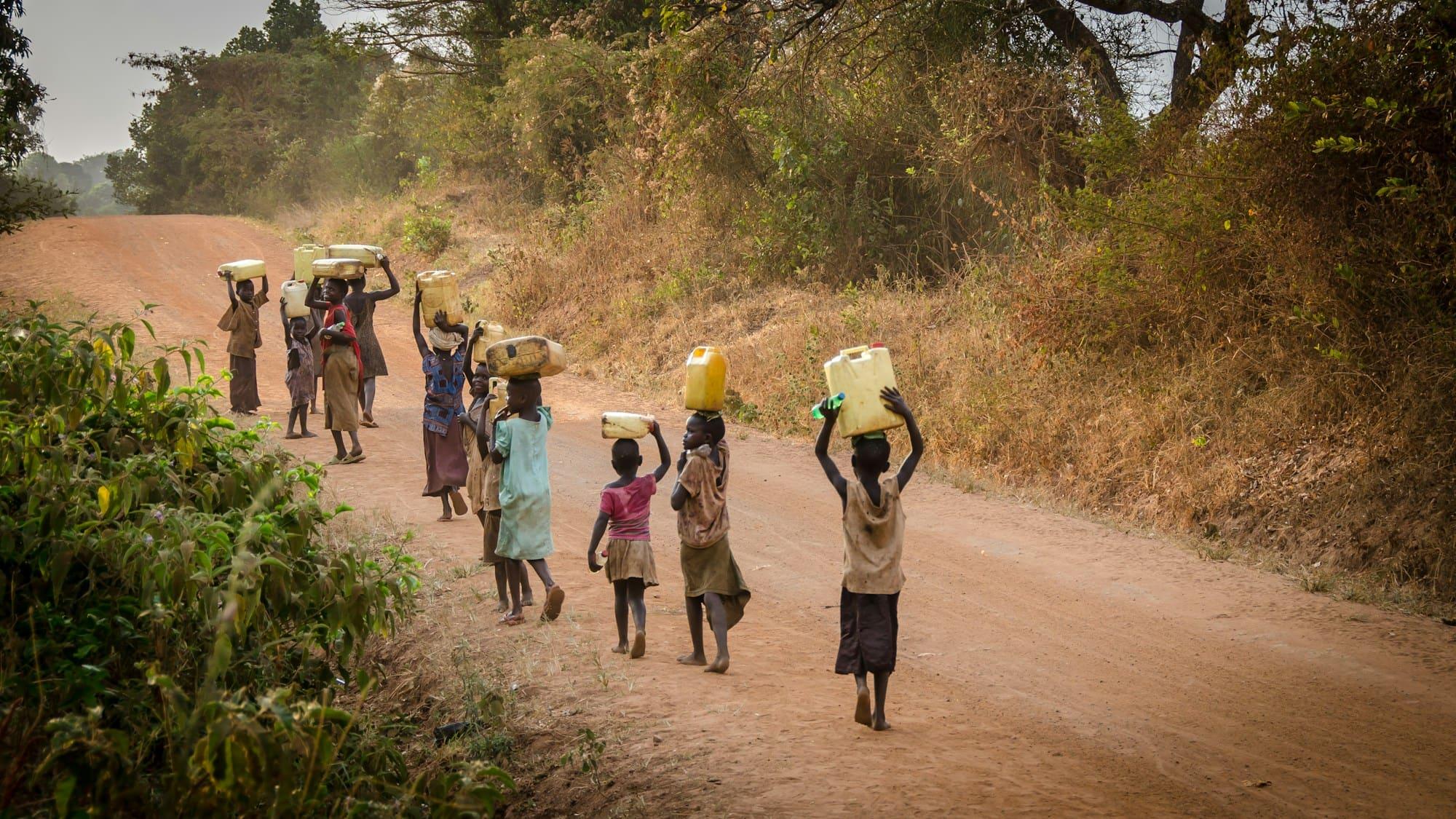 Children and adults walk along a dirt road carrying water containers on their heads